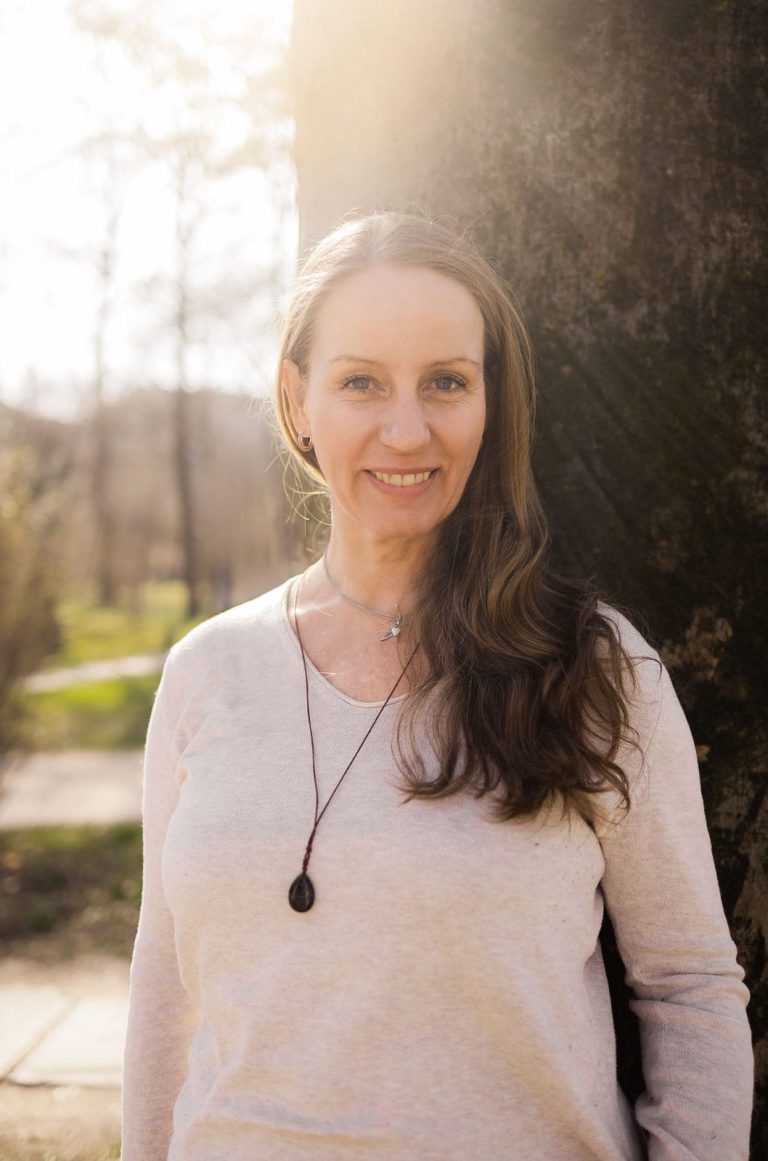 Eine Frau mit langen Haaren lächelt, trägt ein weißes Oberteil und steht in der Natur.