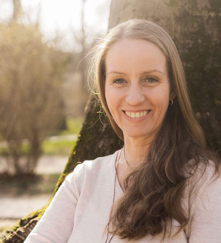 Lächelnde Frau mit langen Haaren, sitzt an einem Baum im Sonnenlicht.